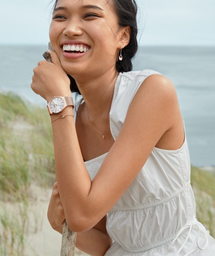 Eine Frau mit einer weißen Bluse, einer Uhr und Schmuck von Skagen. Ein Skagen Set aus Uhr und Armband im Sand.